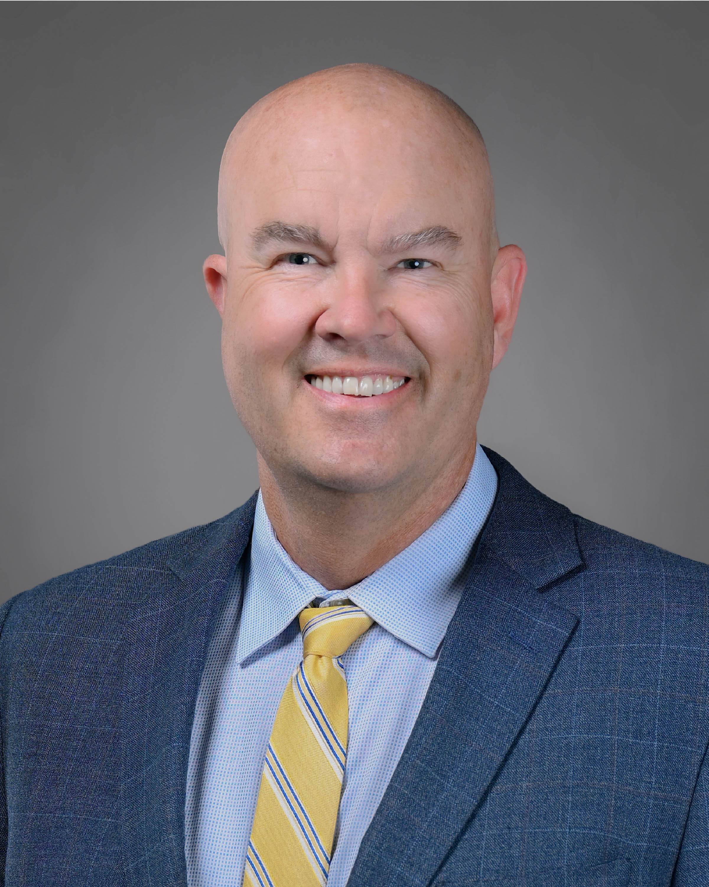 Smling man with a clean shaven head and face with a blue suit and yellow power tie on.